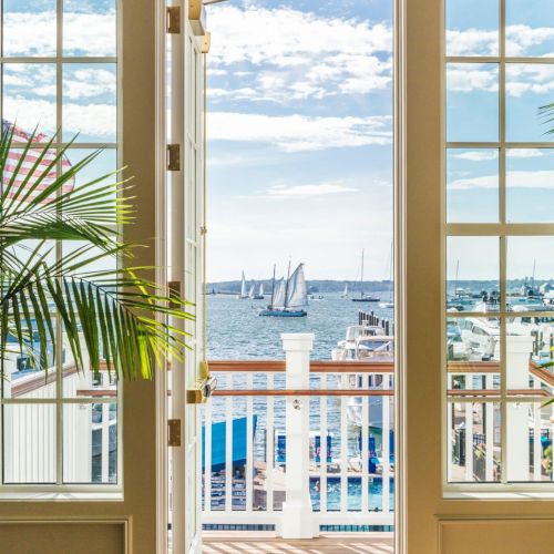 An open door leads to a balcony overlooking a scenic harbor with boats and a clear sky, framed by two large windows and a potted plant.