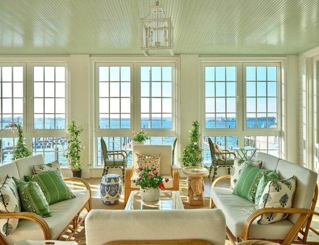 A sunlit living room with large windows, green and white decor, wicker furniture, and a view of water outside, adorned with plants and cushions.