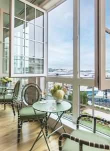 A cozy indoor seating area with a table and chairs by large open windows, offering a scenic view outside. The table has flowers and a drink.