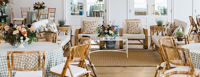 The image shows a well-decorated event space with wicker chairs, round tables with checkered tablecloths, and floral arrangements.