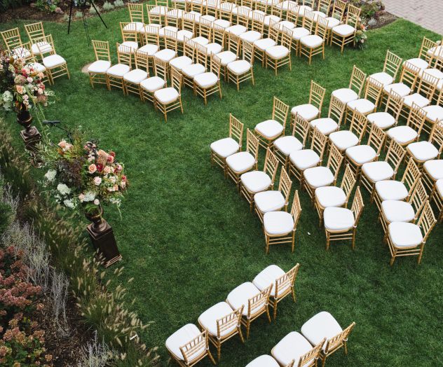 The image shows an outdoor wedding or event setup with rows of light-colored chairs on a green lawn, and some floral arrangements near the chairs.