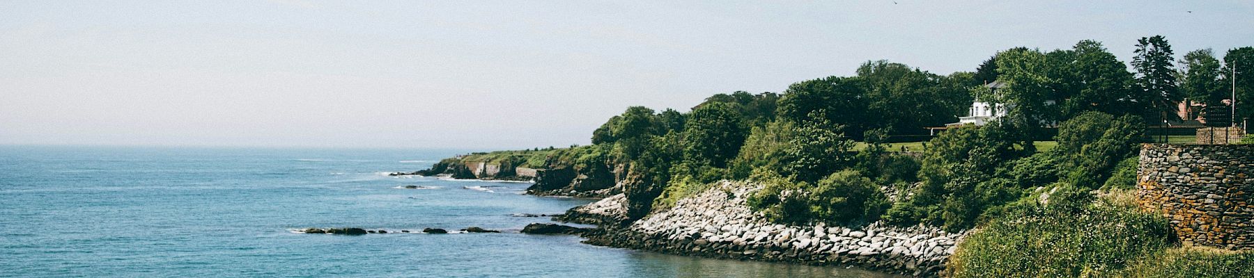 A serene coastal scene with clear skies, cliffs, lush green foliage, and gentle waves breaking against rocks and a distant horizon.