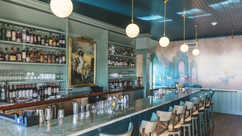 A stylish bar with a long counter, bar stools, shelves of bottles, a painting, globe lights, and a mural on the back wall, creating a cozy ambiance.