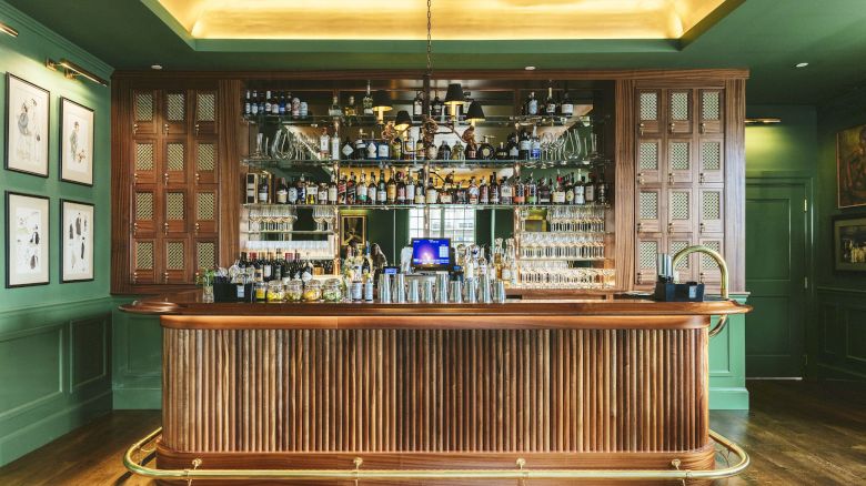 The image shows a well-lit, elegant bar with wooden decor, various bottles, and glasses on display, with green walls and framed pictures hanging.