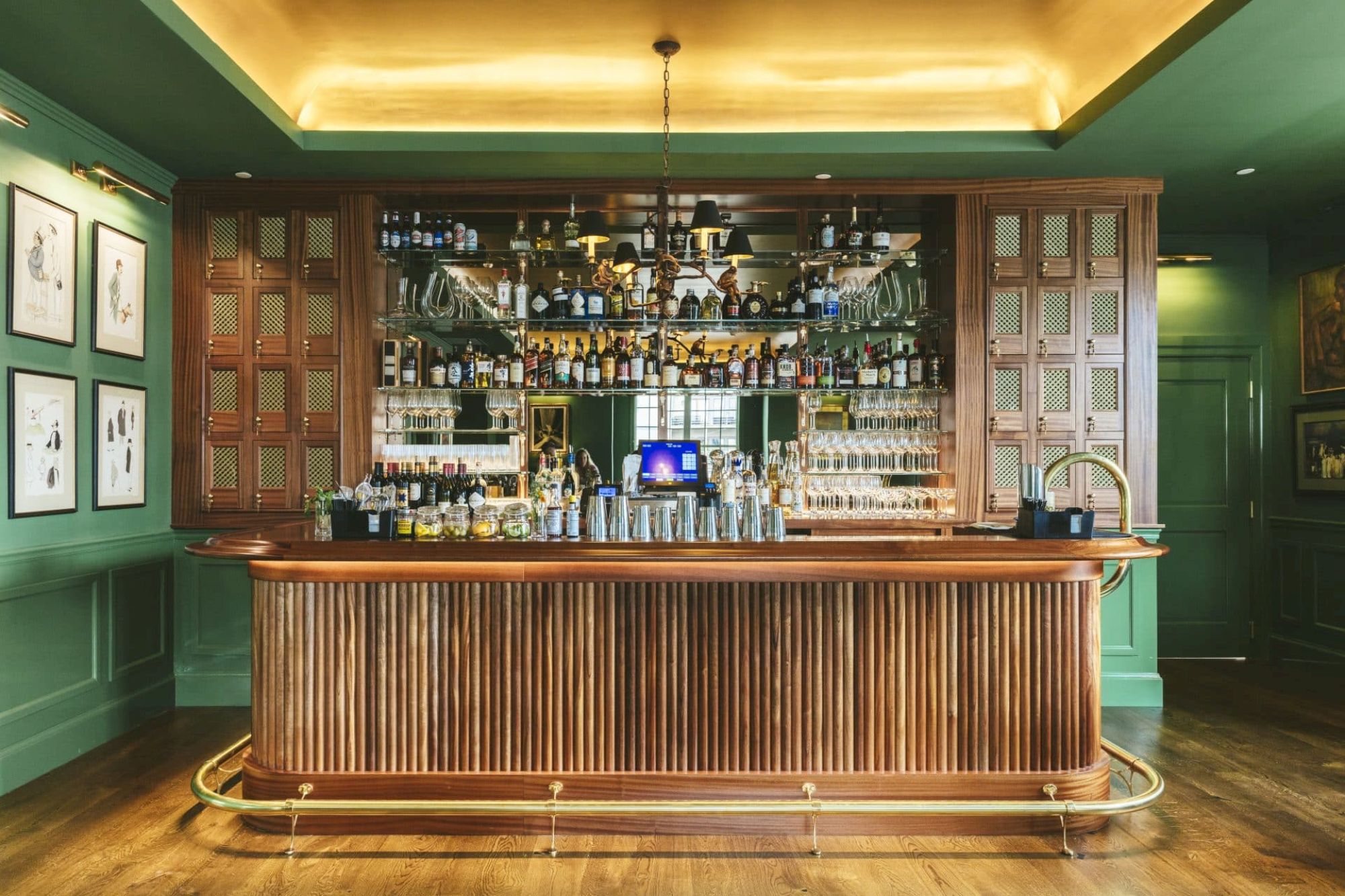 A well-stocked, elegant bar with wood paneling, under warm lighting, features a variety of liquor bottles behind; framed art hangs on the green walls.