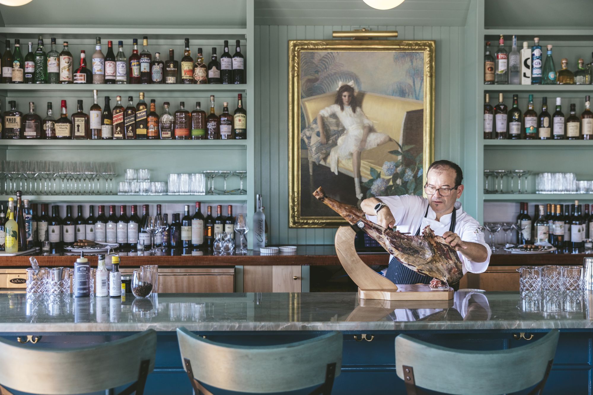 A man is slicing prosciutto in a well-stocked bar with a painting of a dog in the background. The bar has various bottles and glassware on display.