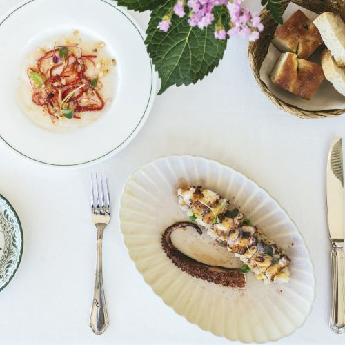 A white table setup with a decorative plate, a seafood appetizer, a dish of bread pieces, a fork and knife, and a flower centerpiece.