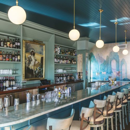 A stylish bar with shelves of liquor bottles, glassware, a marble countertop, pendant lights, a large painting, and a scenic mural on the wall.