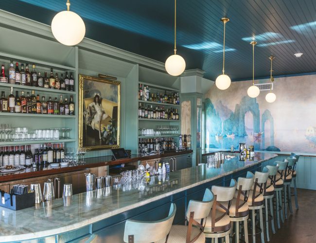 A stylish bar with shelves of liquor bottles, glassware, a marble countertop, pendant lights, a large painting, and a scenic mural on the wall.