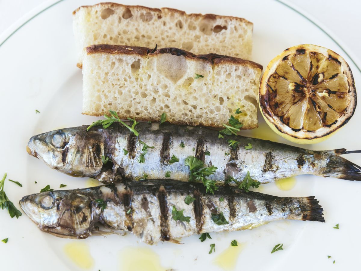 The image shows a plate with two grilled fish, two slices of bread, and a grilled lemon half, garnished with herbs and a drizzle of olive oil.