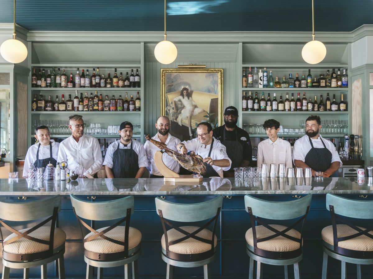 A group of people, likely restaurant staff, stands behind a bar counter. The background includes shelves with bottles and a painting.