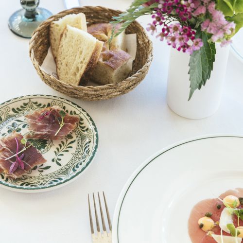 The image shows a table setting with plates of food, a basket of bread, a fork, and a vase containing flowers. The plates hold delicately arranged dishes.