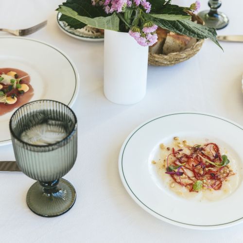 A table setting featuring three elegant plates of food, a drink, a vase with flowers, and cutlery arranged neatly on a white tablecloth.