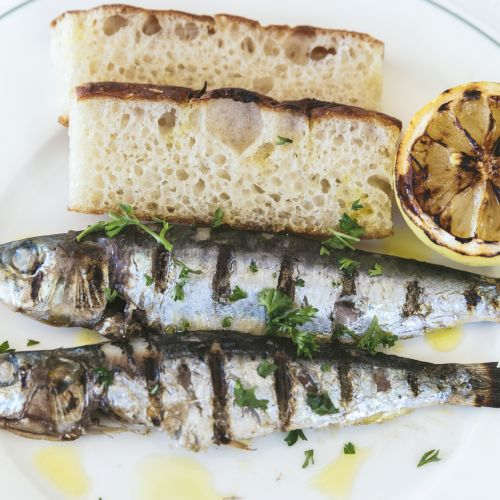 The image shows two grilled fish, garnished with herbs, accompanied by toasted bread slices and a grilled lemon half on a white plate.