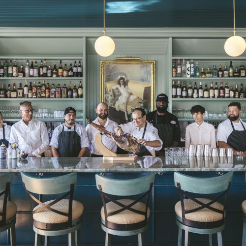 A group of restaurant staff stand behind a bar counter, with shelves of bottles and glasses behind them, and a painting on the wall.