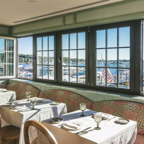 A bright, cozy restaurant with large windows overlooking a marina filled with boats. Tables are set with white linens and glassware.