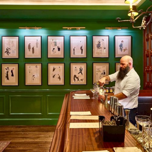 A bartender with a beard is mixing drinks behind a wooden bar in a green-painted room with framed pictures on the wall in front of him.