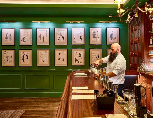 A bartender with a beard is mixing drinks behind a wooden bar in a green-painted room with framed pictures on the wall in front of him.