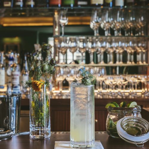 A well-stocked bar with various bottles and glasses, featuring a flower vase, a tall cocktail, and fresh garnishes on the counter.