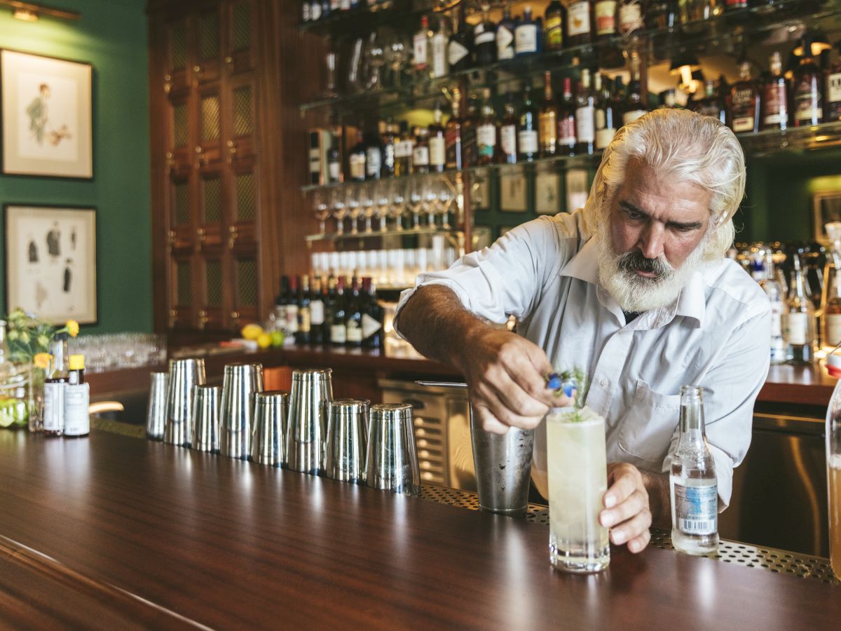 A bartender with grey hair prepares a drink at a bar with a variety of bottles and a neatly organized setup on the counter.