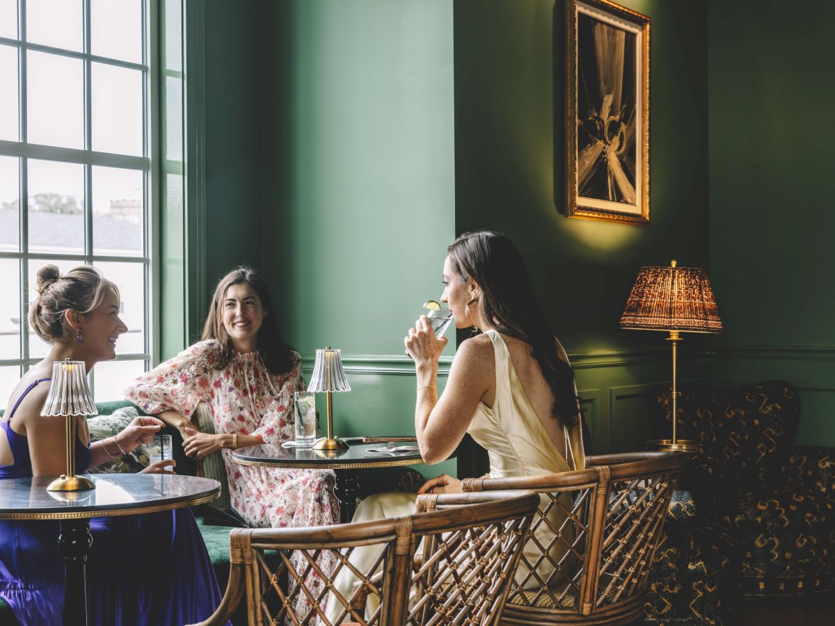 Three women are sitting in a green-painted room, chatting and drinking by the window. The room is elegantly decorated with vintage furniture.